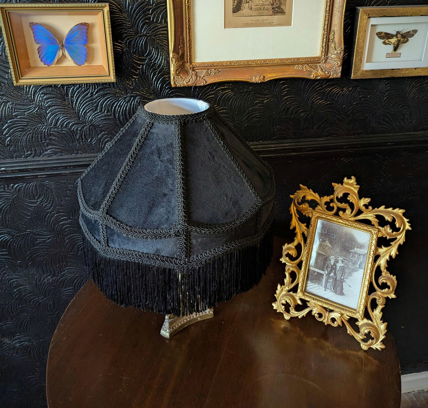 Black lampshade with fringe on a wooden surface, framed pictures in the background