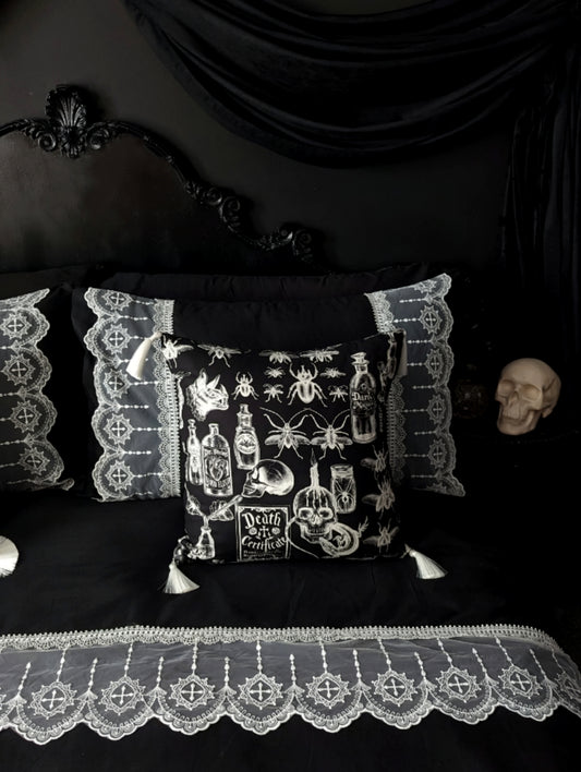 Mourning Parlour black lace bedding from Joely Ball Home. Wide bands of white scalloped lace adorn a  black pillowcase and duvet cover on a black Rococco bed. The wall is black with black drapery and a skull sits on the bedside.