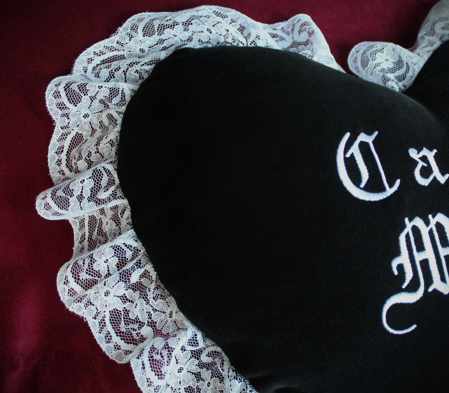 A close up of a black velvet cushion with a white lace trim and letters embroidered in white.