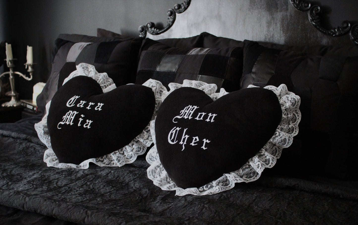 A Gothic bedroom with black cushions and a black throw on a bed with two black heart shaped cushions trimmed in white lace in the centre. One has "Cara Mia" embroidered on it and the other has "Mon Cher".