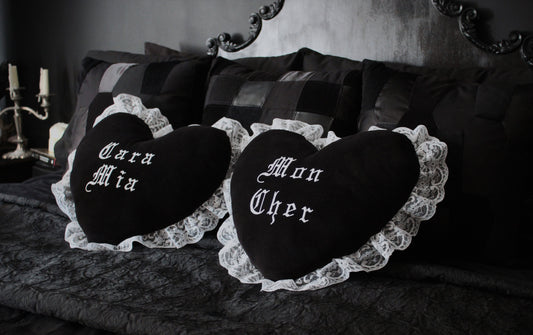 A Gothic bedroom with black cushions and a black throw on a bed with two black heart shaped cushions trimmed in white lace in the centre. One has "Cara Mia" embroidered on it and the other has "Mon Cher".