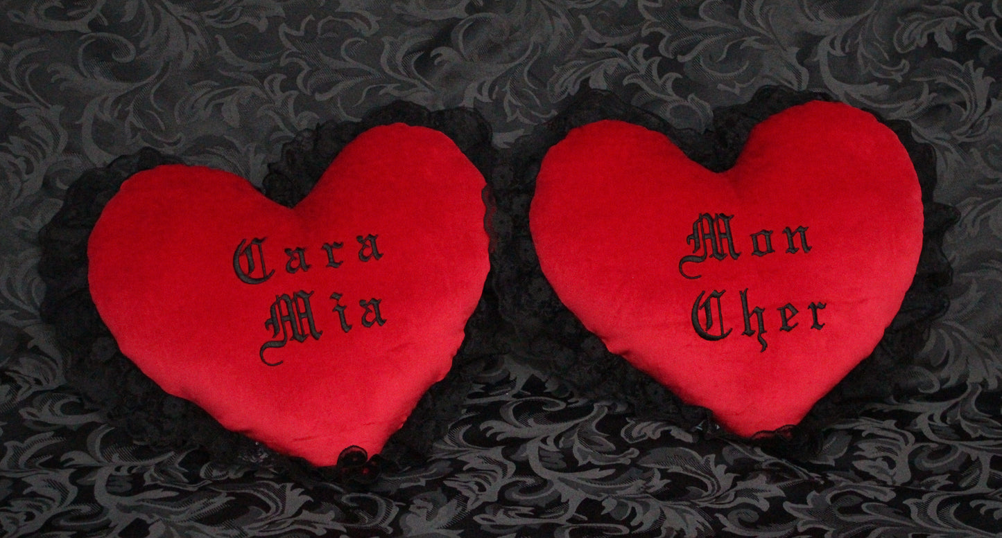 Two red Gothic heart shaped velvet cushions with black lace trim, that say Mon Cher and Cara Mia.