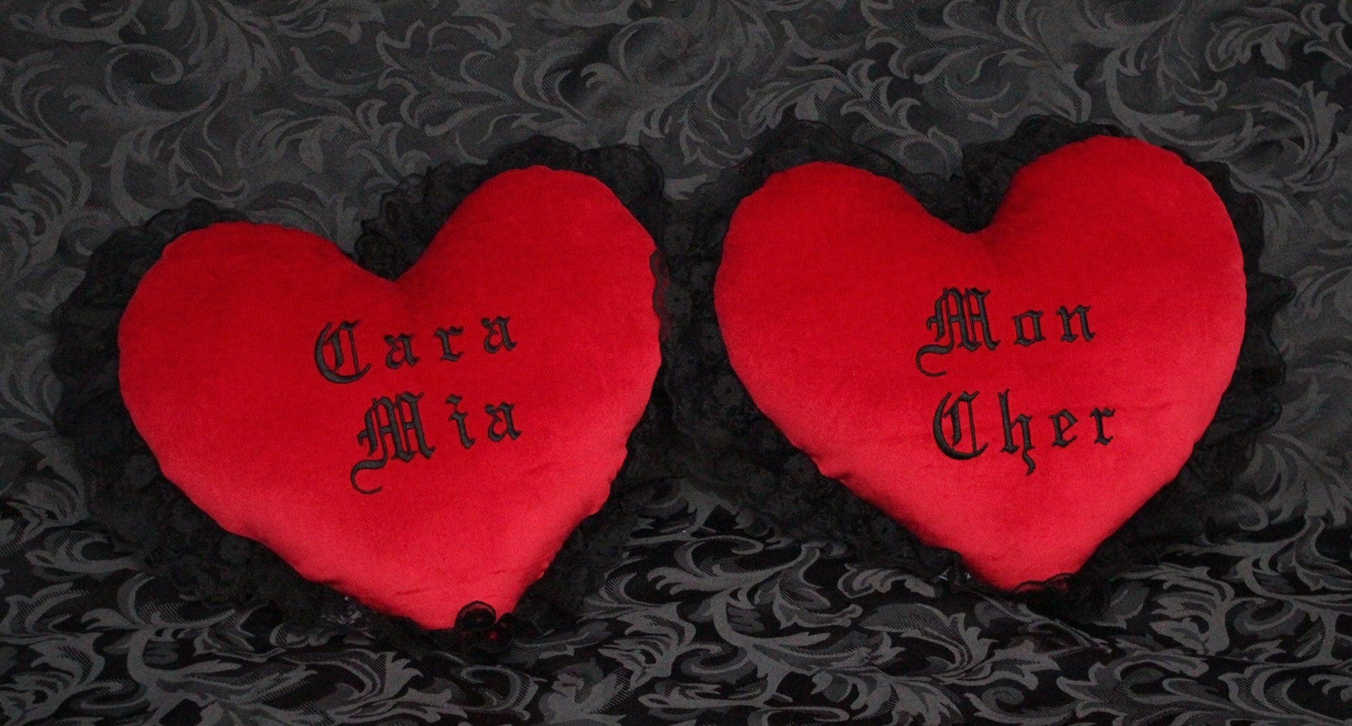 Two red Gothic heart shaped velvet cushions with black lace trim, that say Mon Cher and Cara Mia.