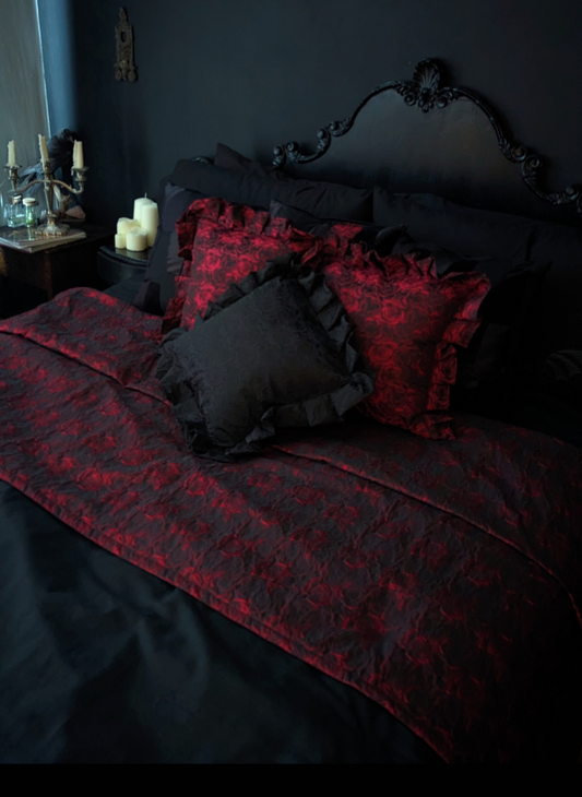 A luxurious red and black bed throw with a floral pattern, displayed on a bed in a dimly lit room, accompanied by matching pillows and candlelight.
