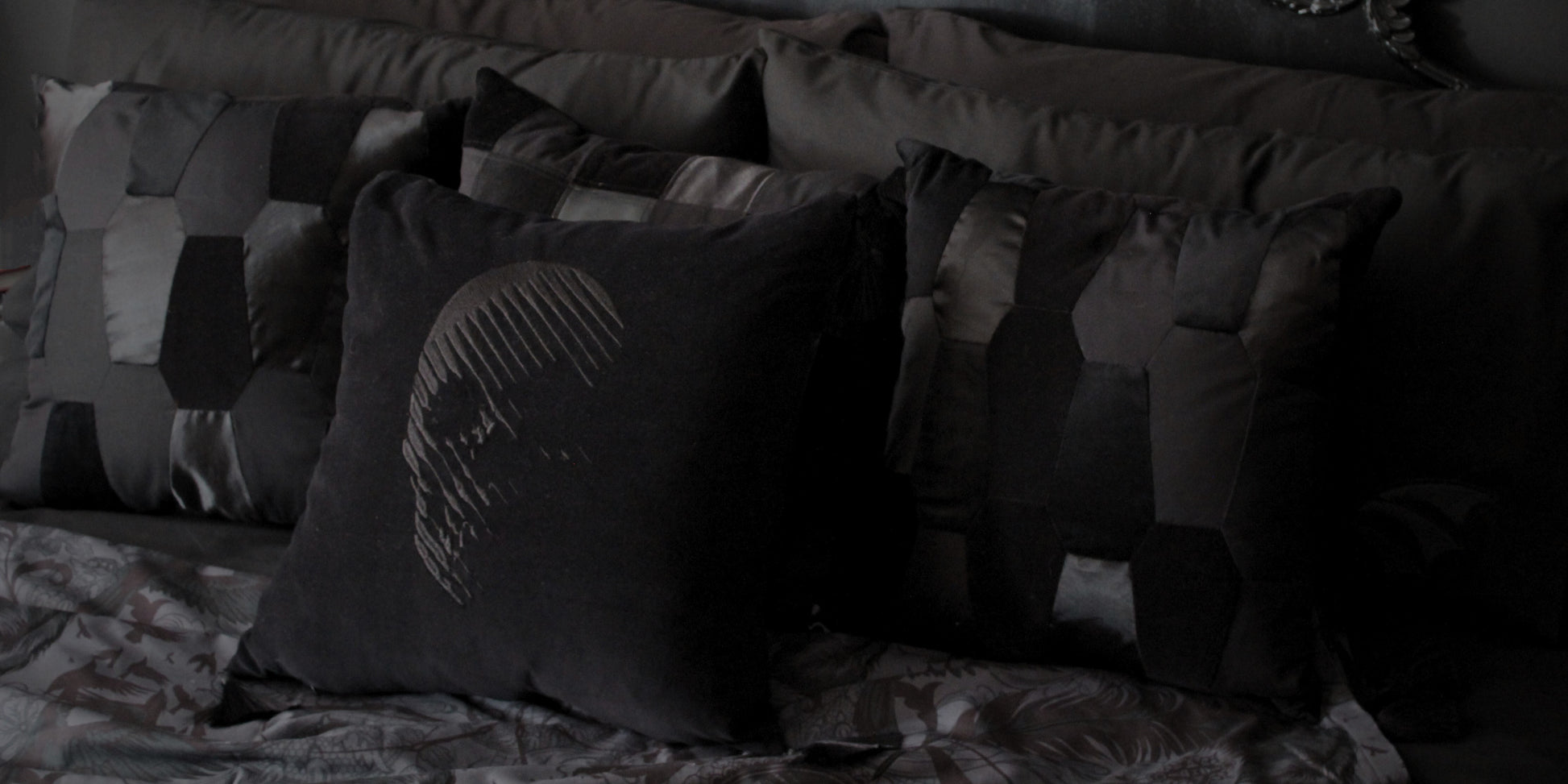 Three black patchwork cushions sit in front of pillows, with an all black cushion with a skull embroidered on it in front.