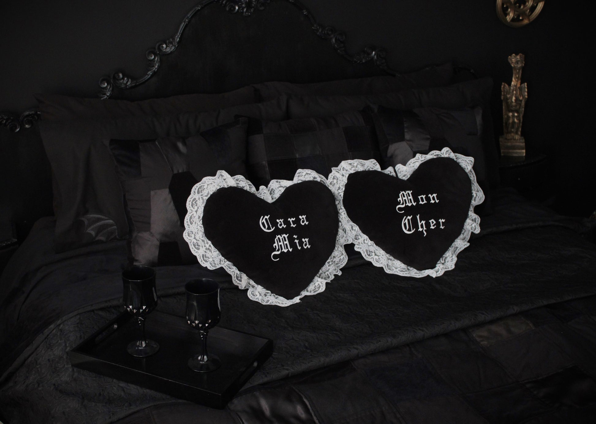 A black bedroom with black walls, black headboard, black patchwork cushions and layers of black throws on the bed. Two black heart cushions with white lace are centre of the bed, one saying Mon Cher and the other Cara Mia. A gargoyle proudly sits in the background and a tray with black wine glasses rests on the bed.