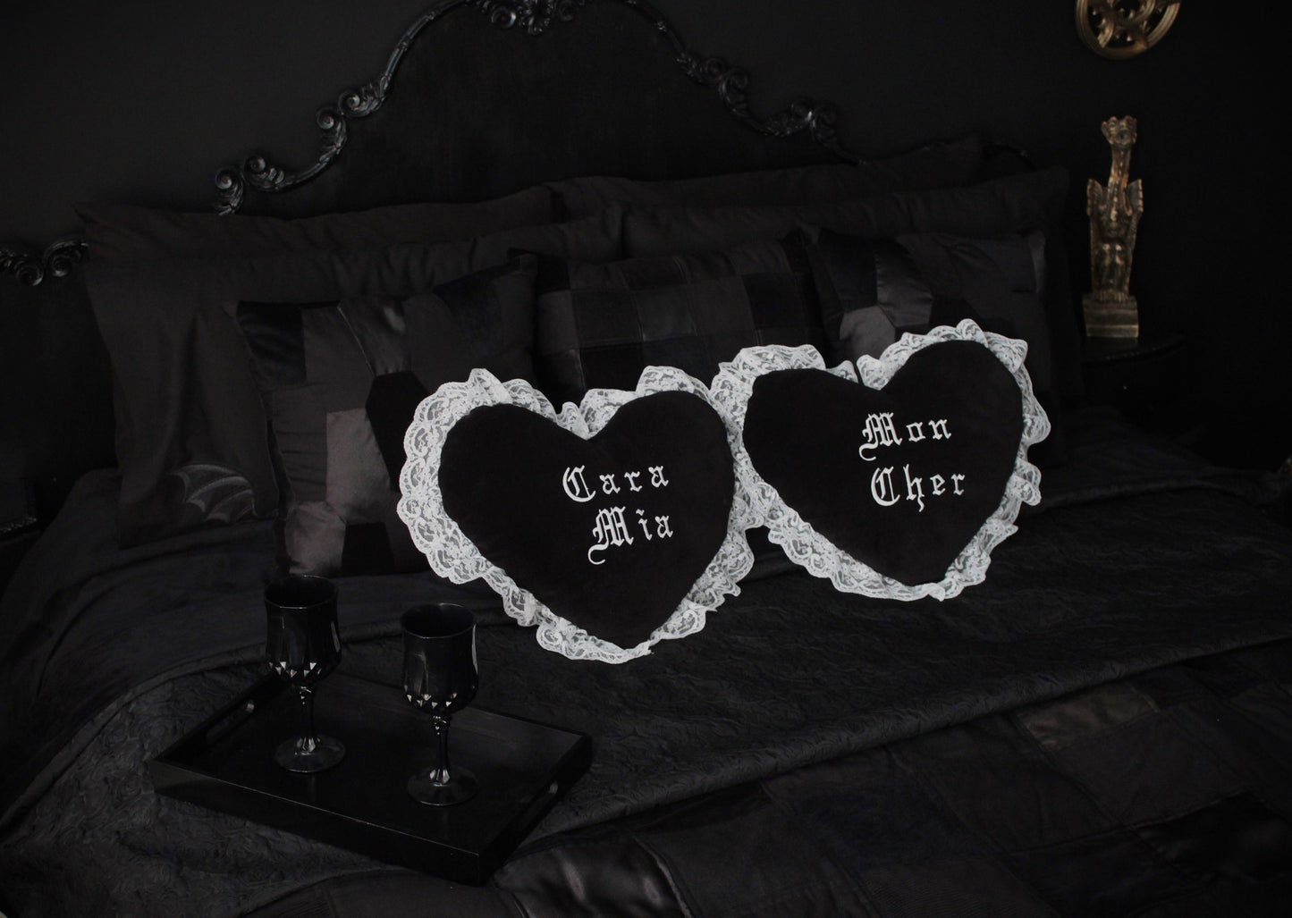 A black bedroom; black walls, black headboard, black patchwork cushions and layers of black throws on the bed. Two cushions sit centre, both black hearts surrounded by white lace, one saying Mon Cher and the other Cara Mia. A gargoyle proudly sits in the background.