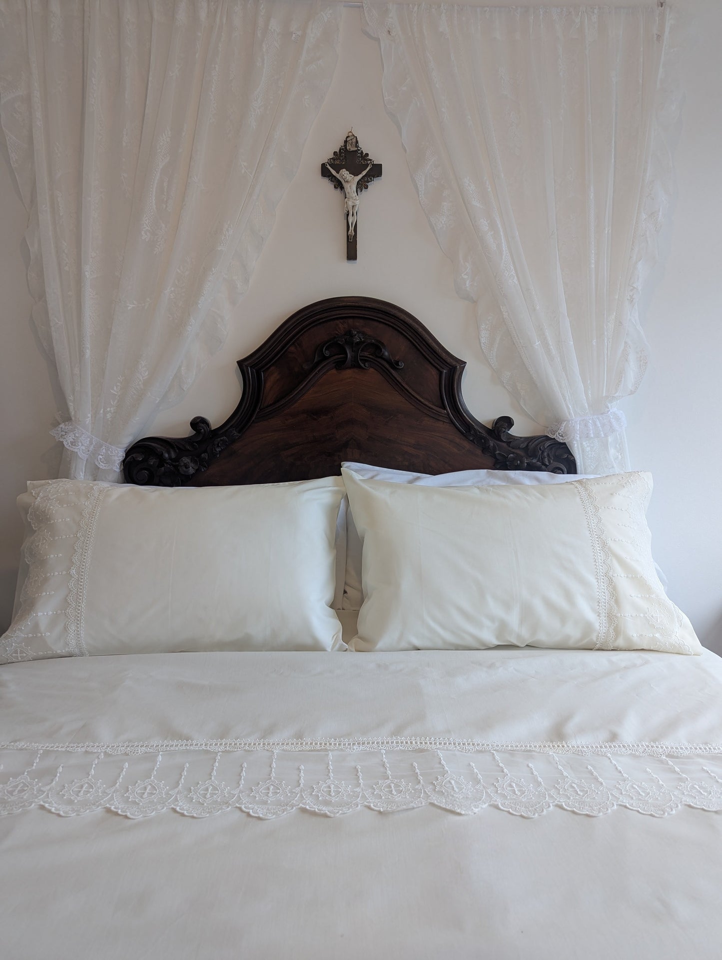 Bed with lace-trimmed bedding and wooden headboard with white wall drapes and a crucifix on the wall behind.