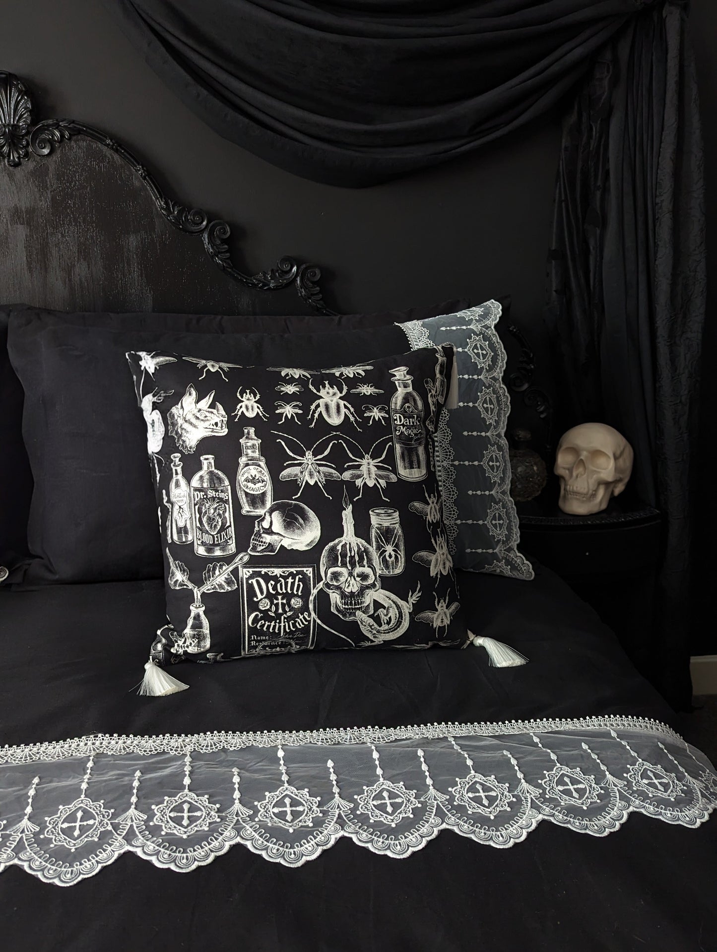 Decorative pillow with skulls and insects design on a black bed with lace trim.