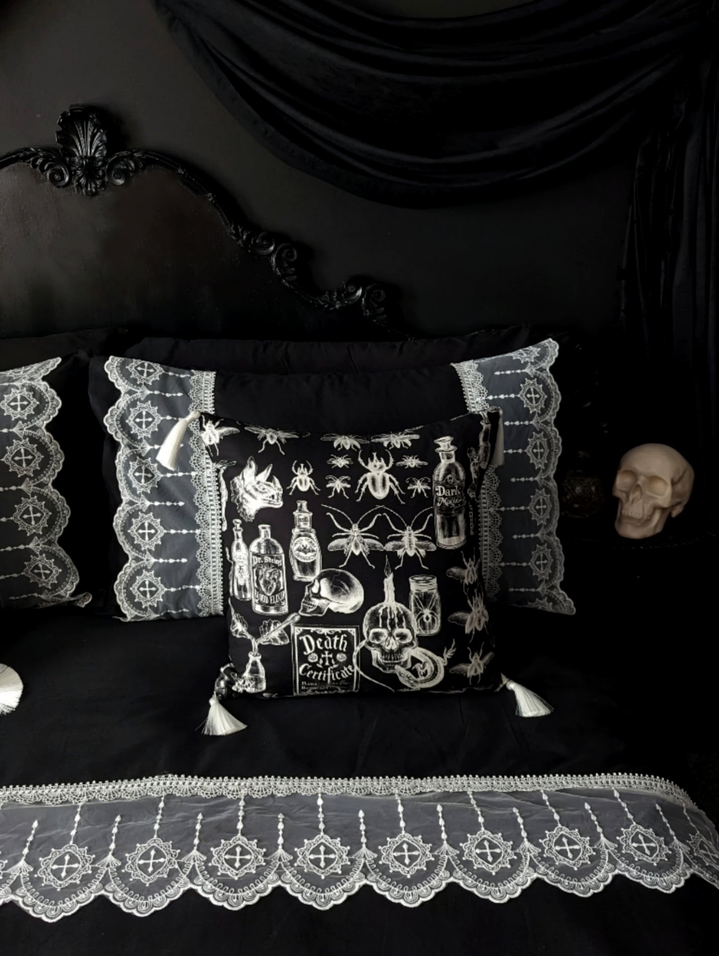 'Mourning Parlour' black lace bedding from Joely Ball Home. White scalloped lace on a black duvet cover and pillowcases on a black Rococo bed. There is a square black cushion with skulls and beetles in white and white tassels. The wall is black with black drapery and a skull sits on the bedside. 