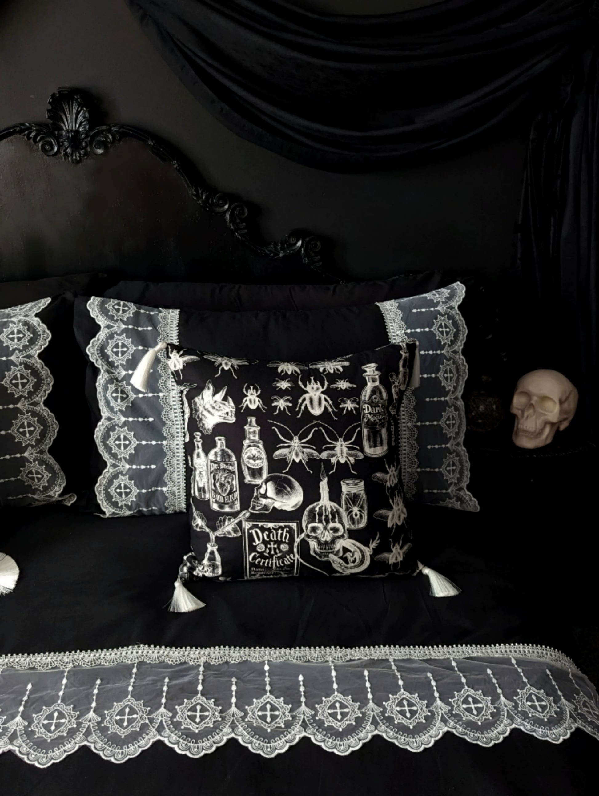 'Mourning Parlour' black lace bedding from Joely Ball Home. White scalloped lace on a black duvet cover and pillowcases on a black Rococo bed. There is a square black cushion with skulls and beetles in white and white tassels. The wall is black with black drapery and a skull sits on the bedside. 