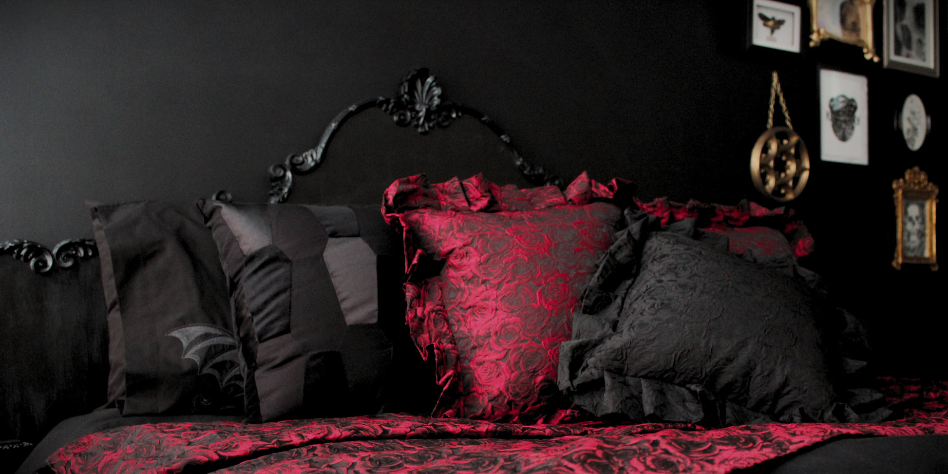A Gothic bedroom, featuring black scatter cushions, ruffled cushions of red roses, a black Rococo headboard on a black wall, with a gallery wall of gold frames in the background.