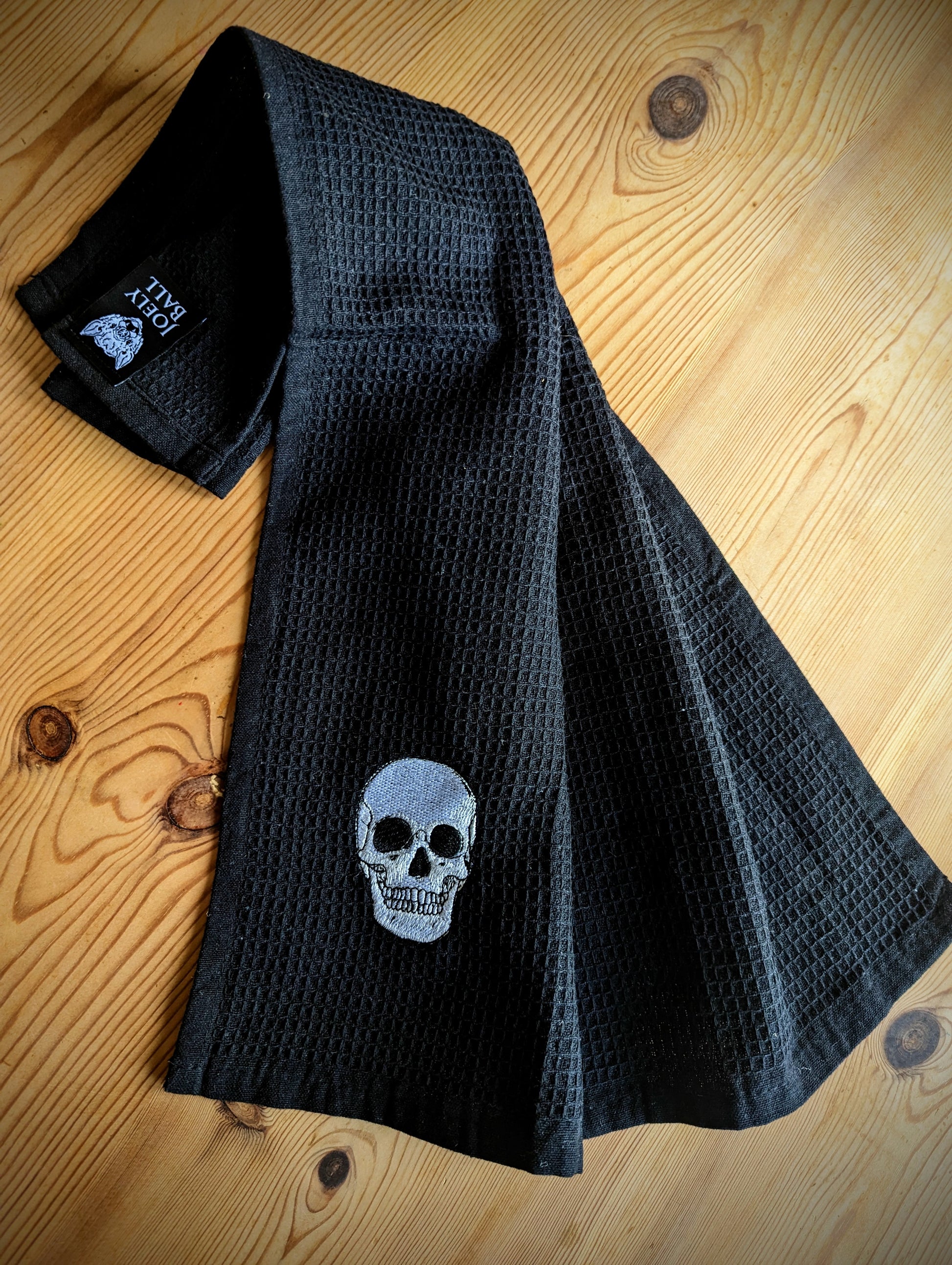 A black cotton waffle tea towel with an embroidered white skull design, laid out on a wooden surface.