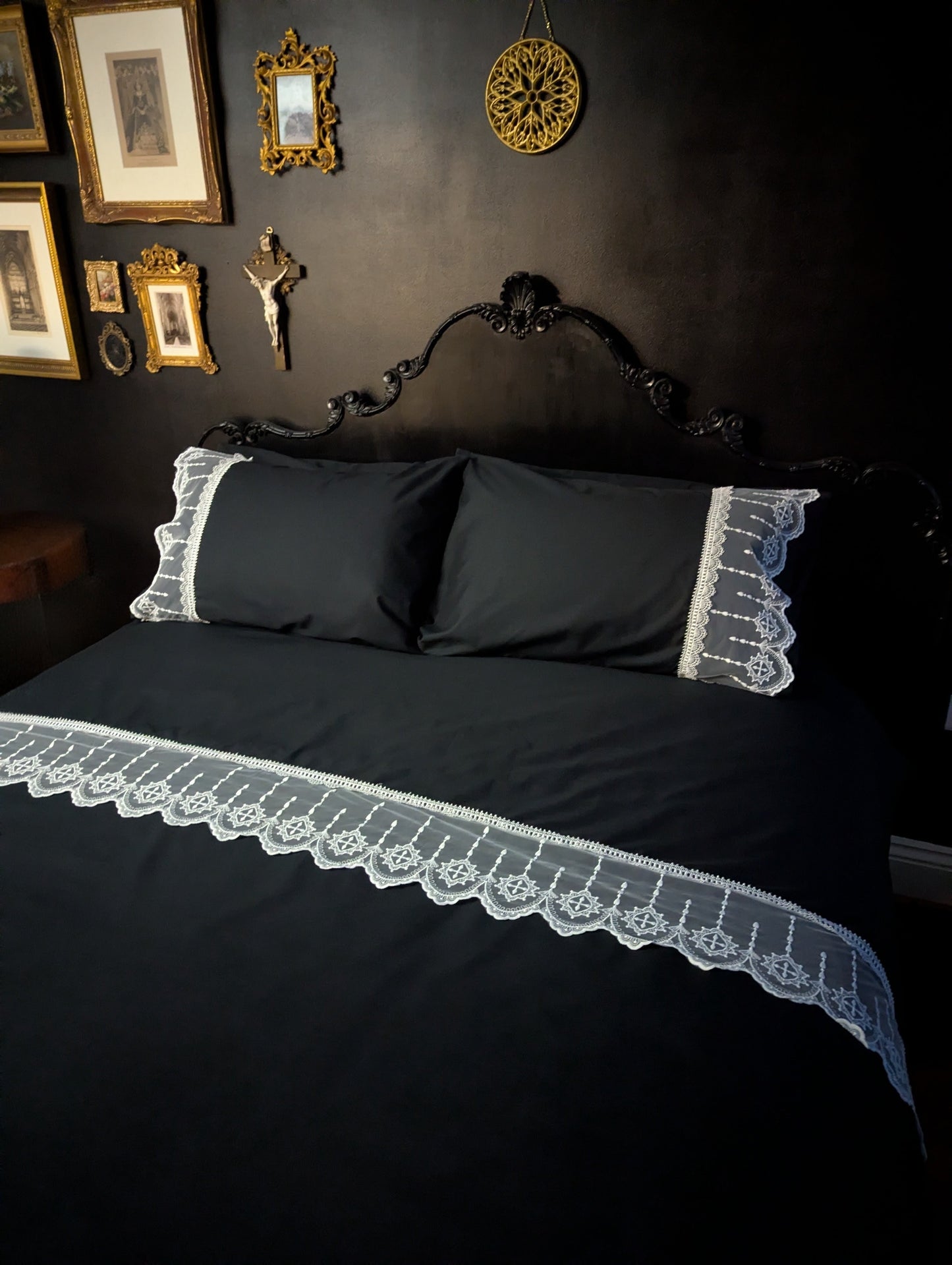 Black bedspread with lace trim on a dark wall with framed pictures.