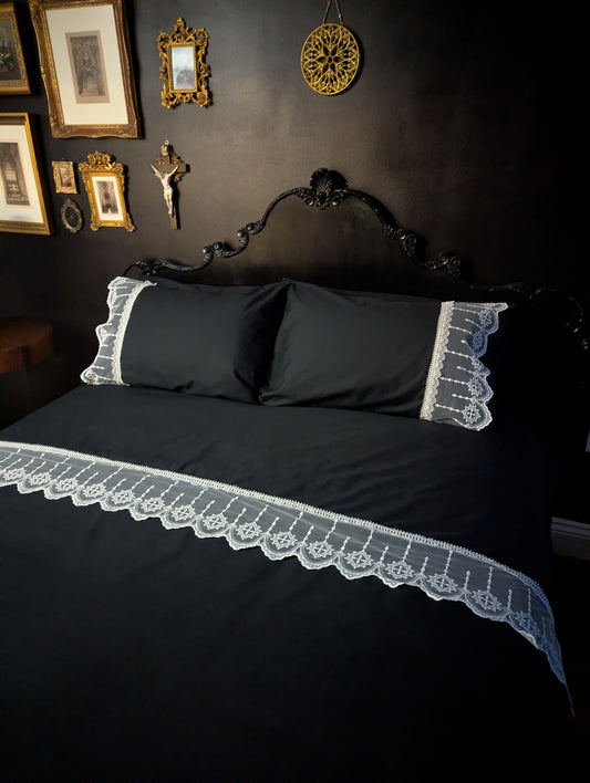 Black bedspread with lace trim on a dark wall with framed pictures.