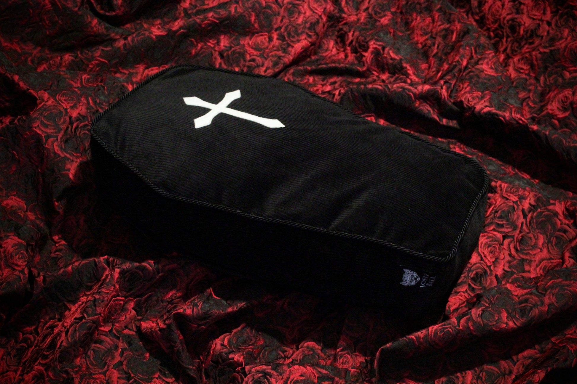 A black corduroy coffin emblazoned with a white cross, sits atop a bed of red roses fabric.