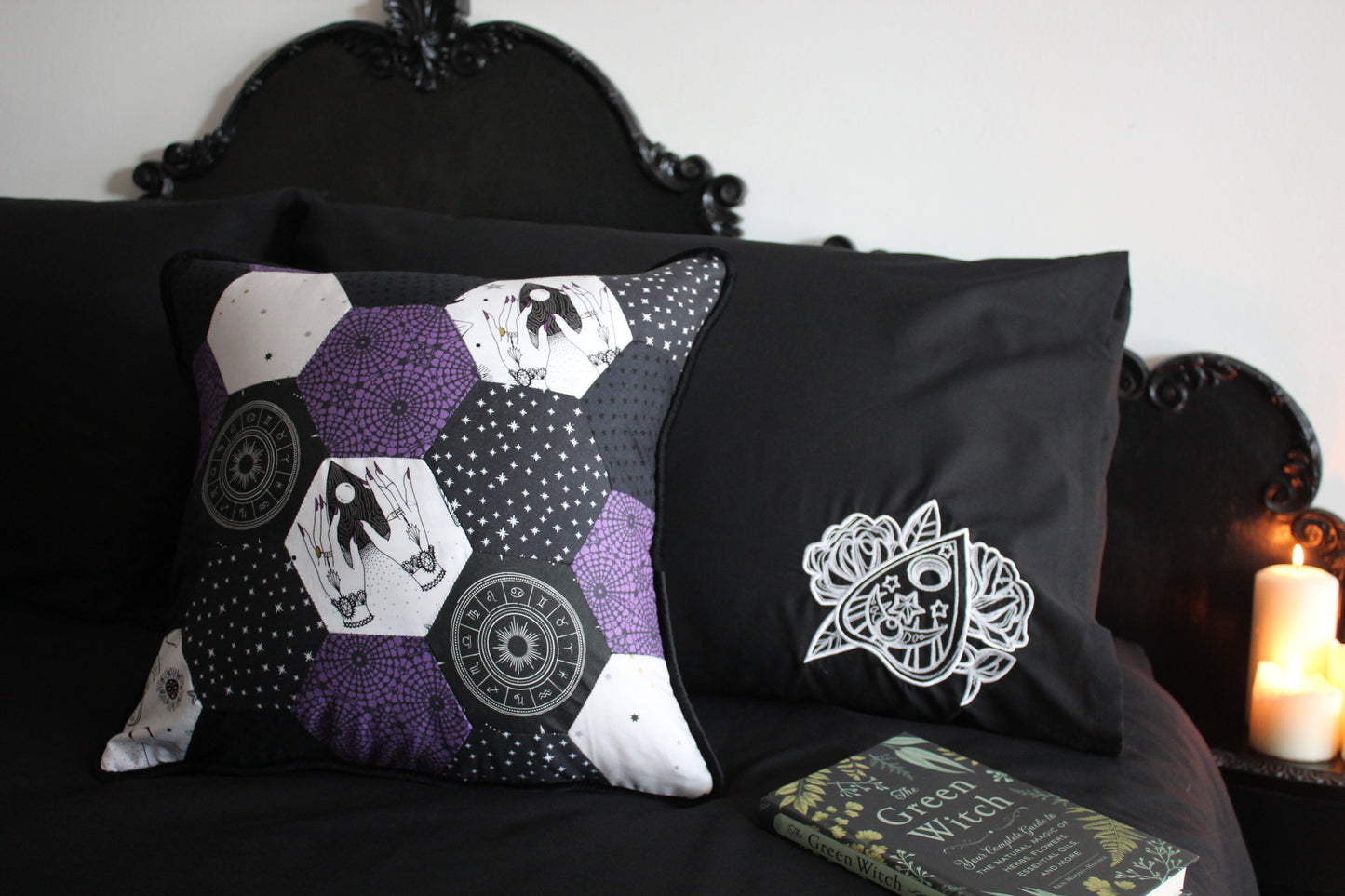 A shot of luxury witchy room decor, featuring an ornate black bed, candles glowing, beside a patchwork cushion in grey, white and purple featuring planchettes and stars. There is a black pillowcase with a white embroidered planchette, with the book The Green Witch in the foreground.