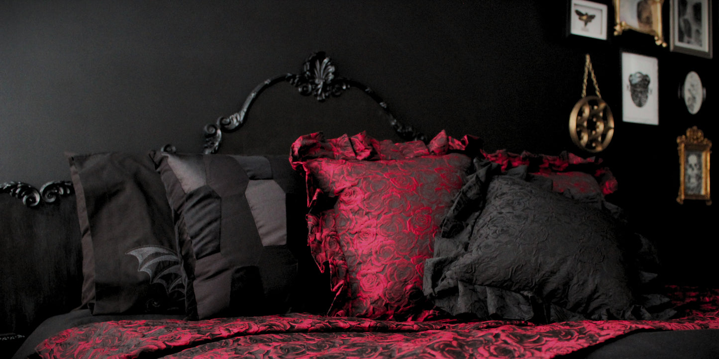 A luxurious Gothic bedroom, featuring black scatter cushions, ruffled cushions with red rose pattern, a black Rococo headboard on a black wall, with a gallery wall of gold frames in the background.