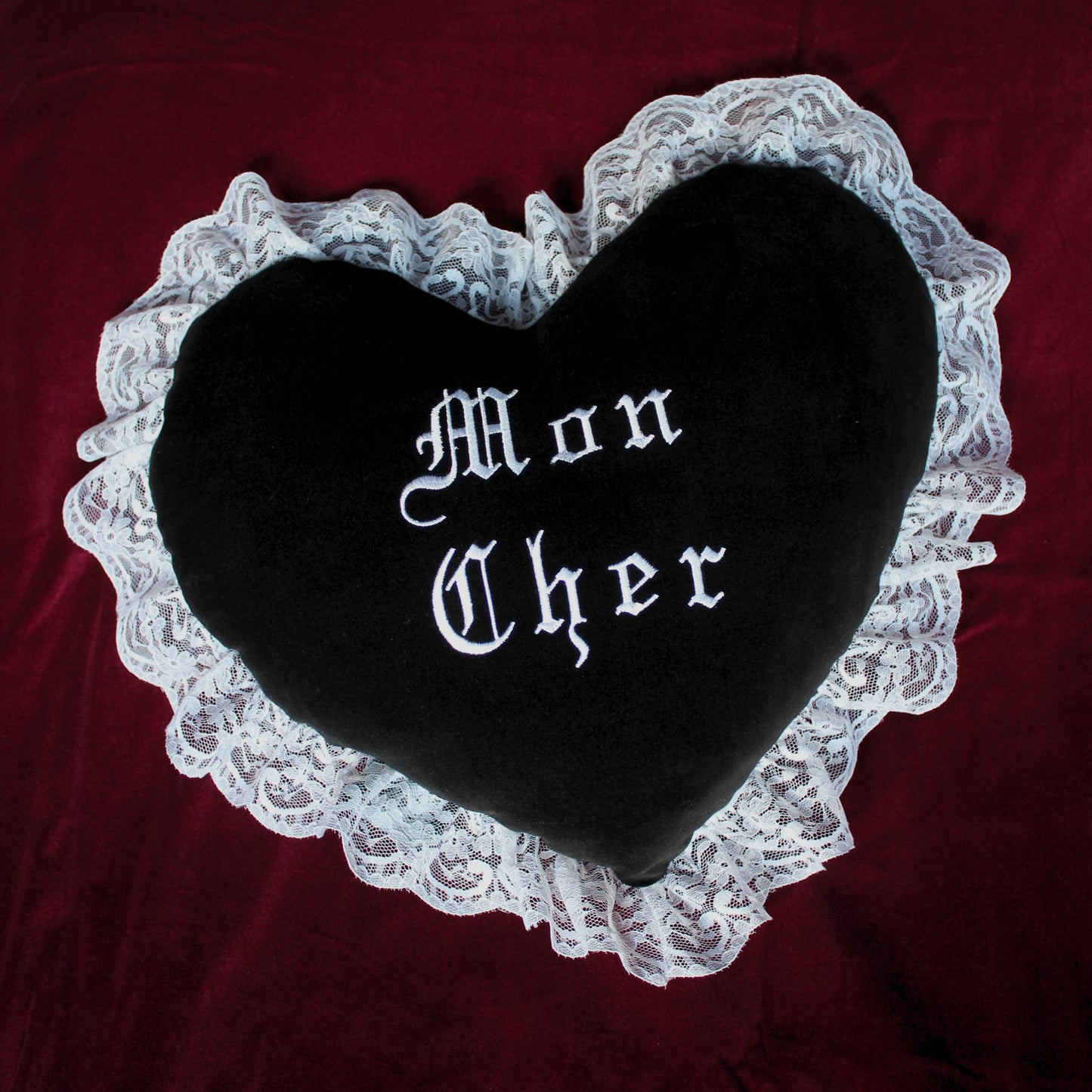 A black velvet heart shaped cushion with a white lace trim. The text Mon Cher is embroidered in the centre, inspired by Morticia and Gomez Addams.