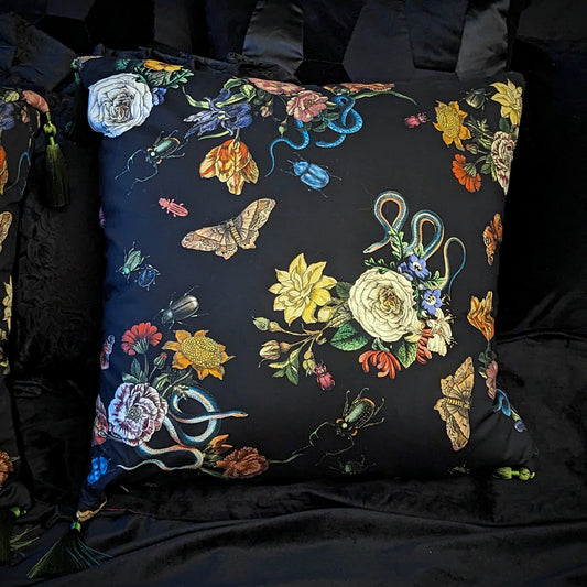 A square cushion with a floral and insect pattern, predominantly dark colored, with green tassels and a black velvet back.