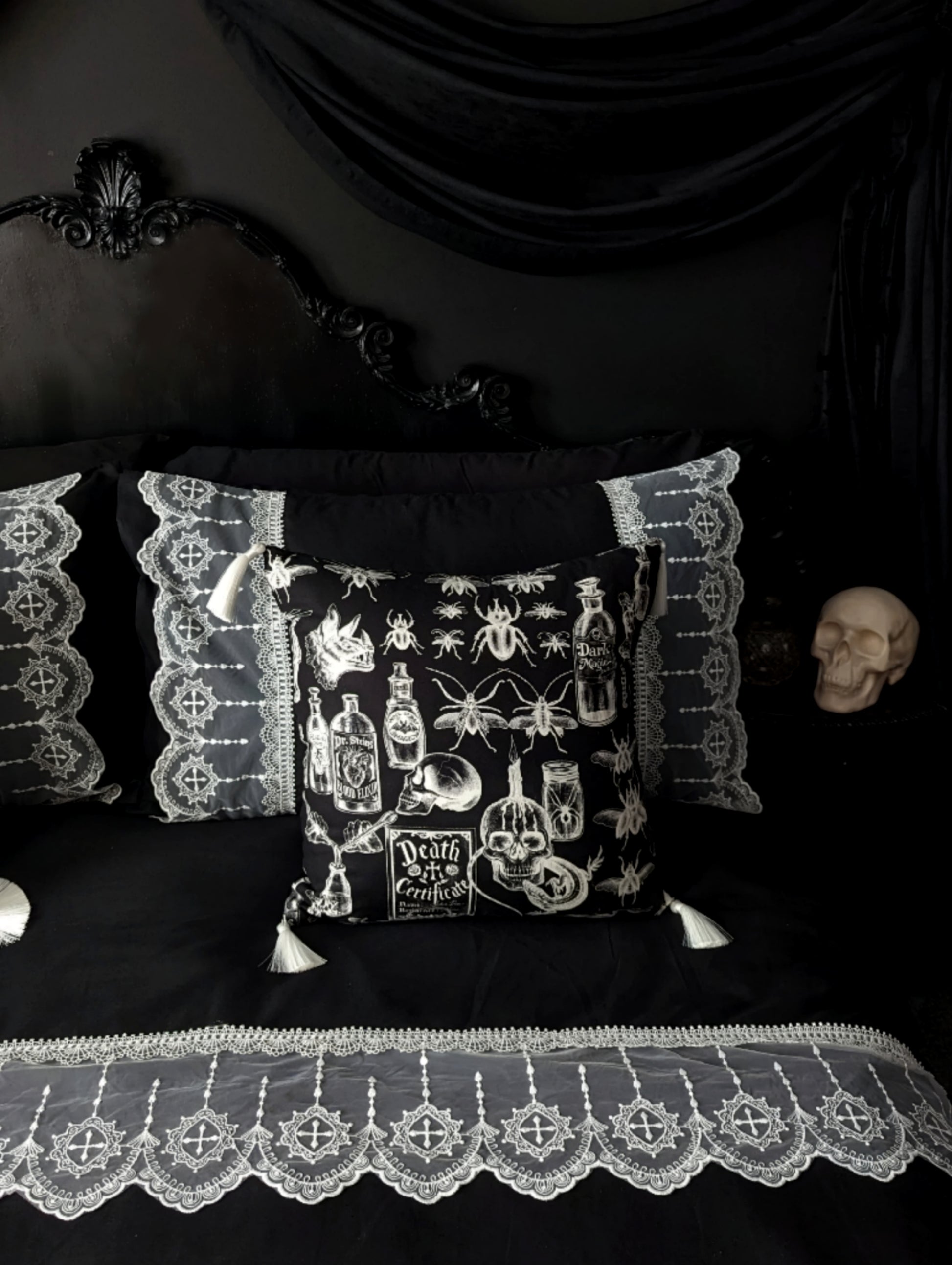 Mourning Parlour black lace bedding from Joely Ball Home. Wide bands of white scalloped lace adorn a  black pillowcase and duvet cover on a black Rococco bed. The wall is black with black drapery and a skull sits on the bedside. 