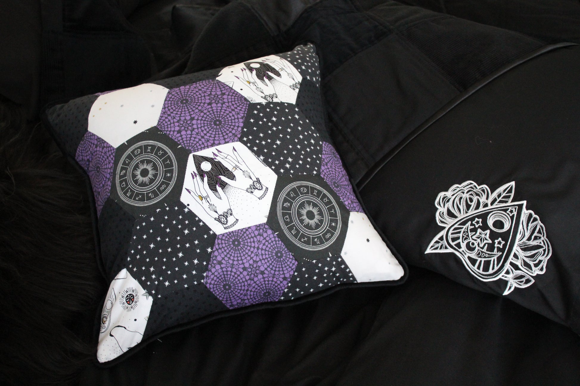 A patchwork cushion made of pink, grey and white hexagons full of mystical images including planchettes and a Zodiac wheel. Beside the cushion there is an embroidered white planchette on a black pillowcase