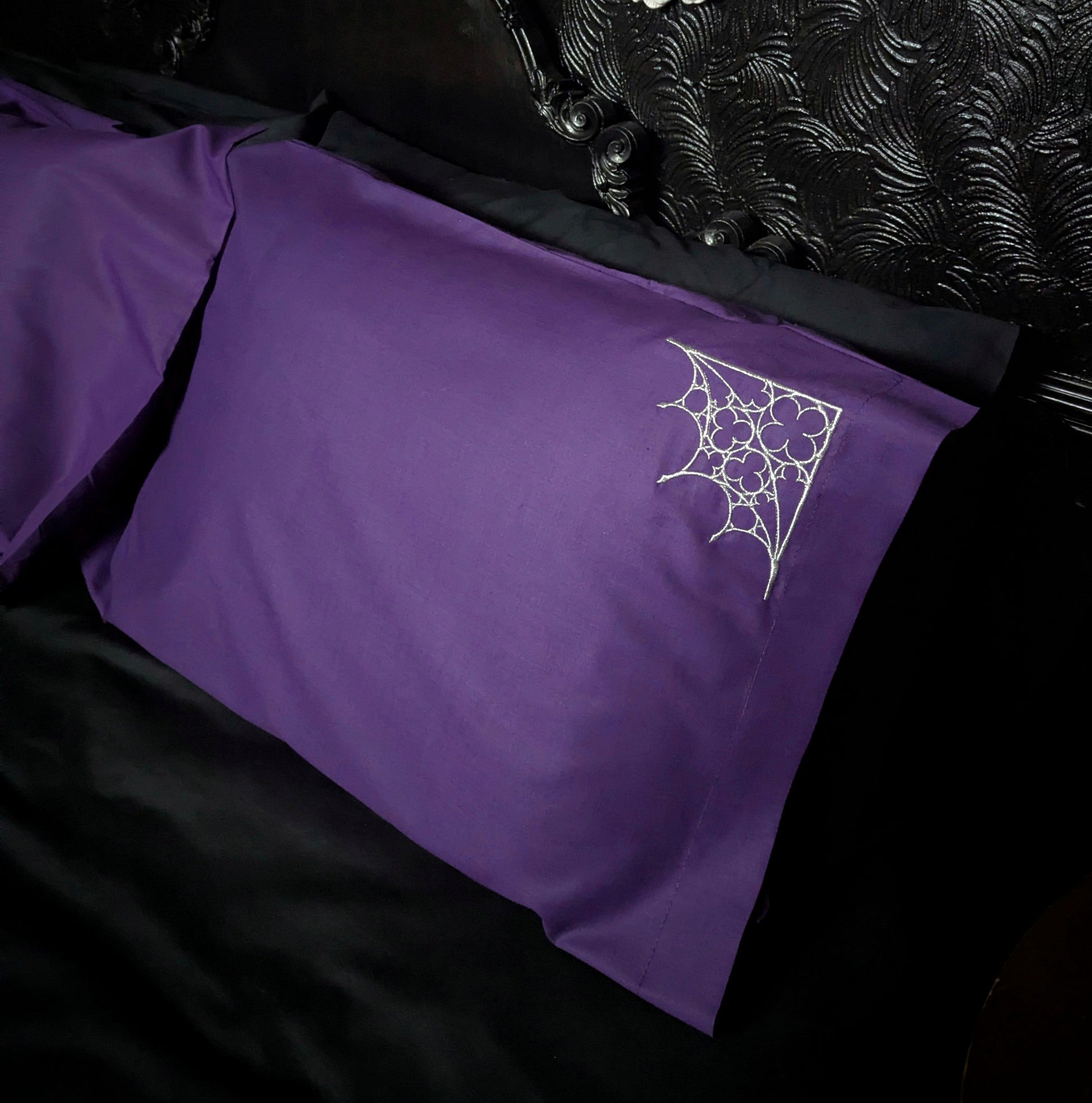 Purple pillowcases with a silver Gothic tracery embroidery on a black bed in front of a black wall 