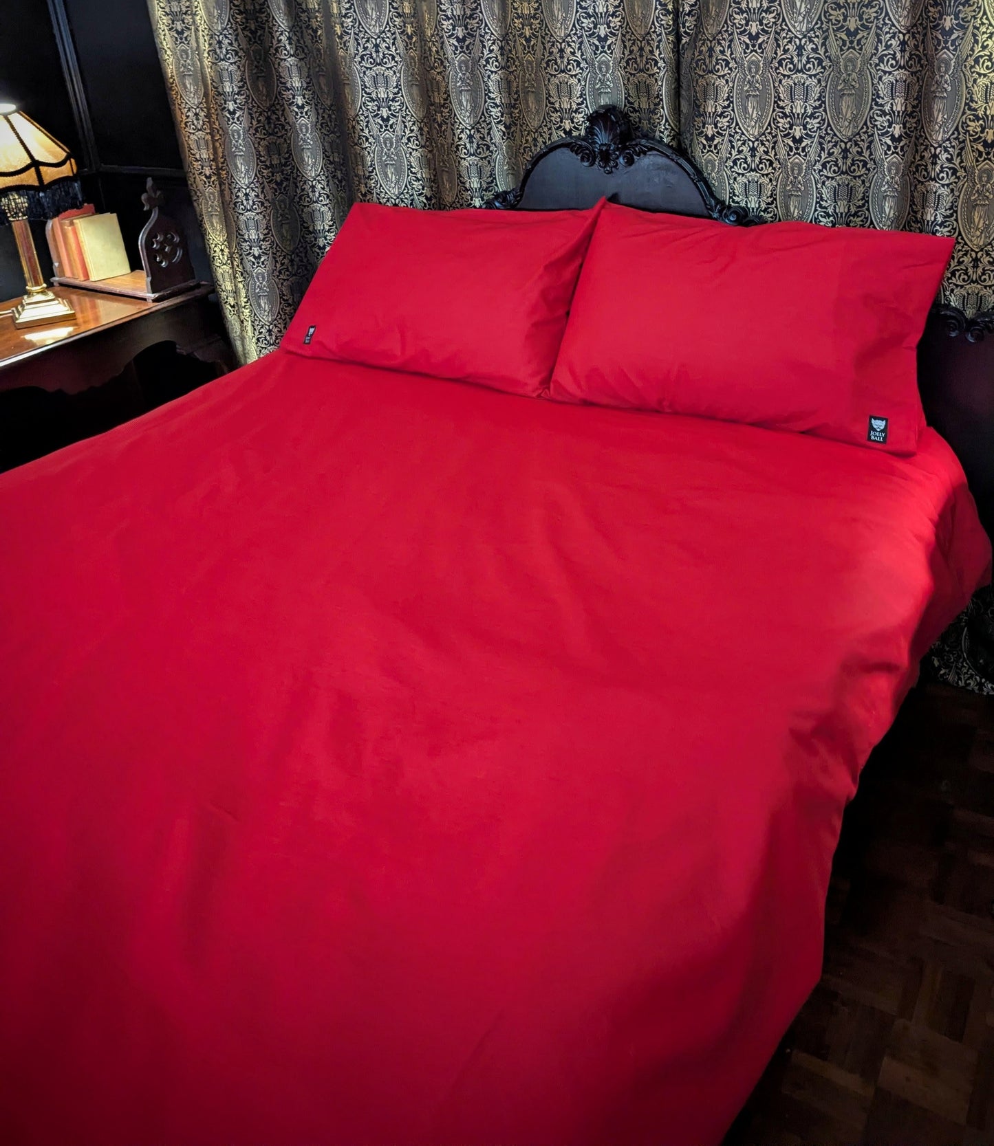 Red bedspread and pillows on a bed with decorative curtains and lamp in the background.