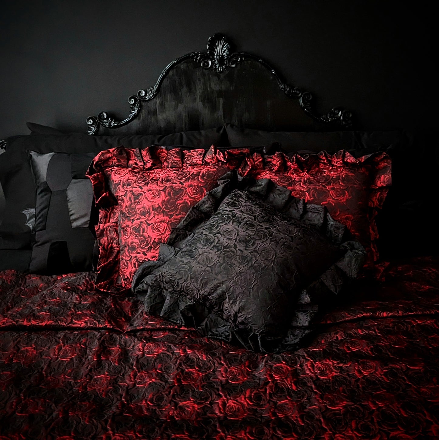 Black and red rose patterned cushions with ruffles around the edge on top of a matching throw in front of a black baroque headboard and black wall.