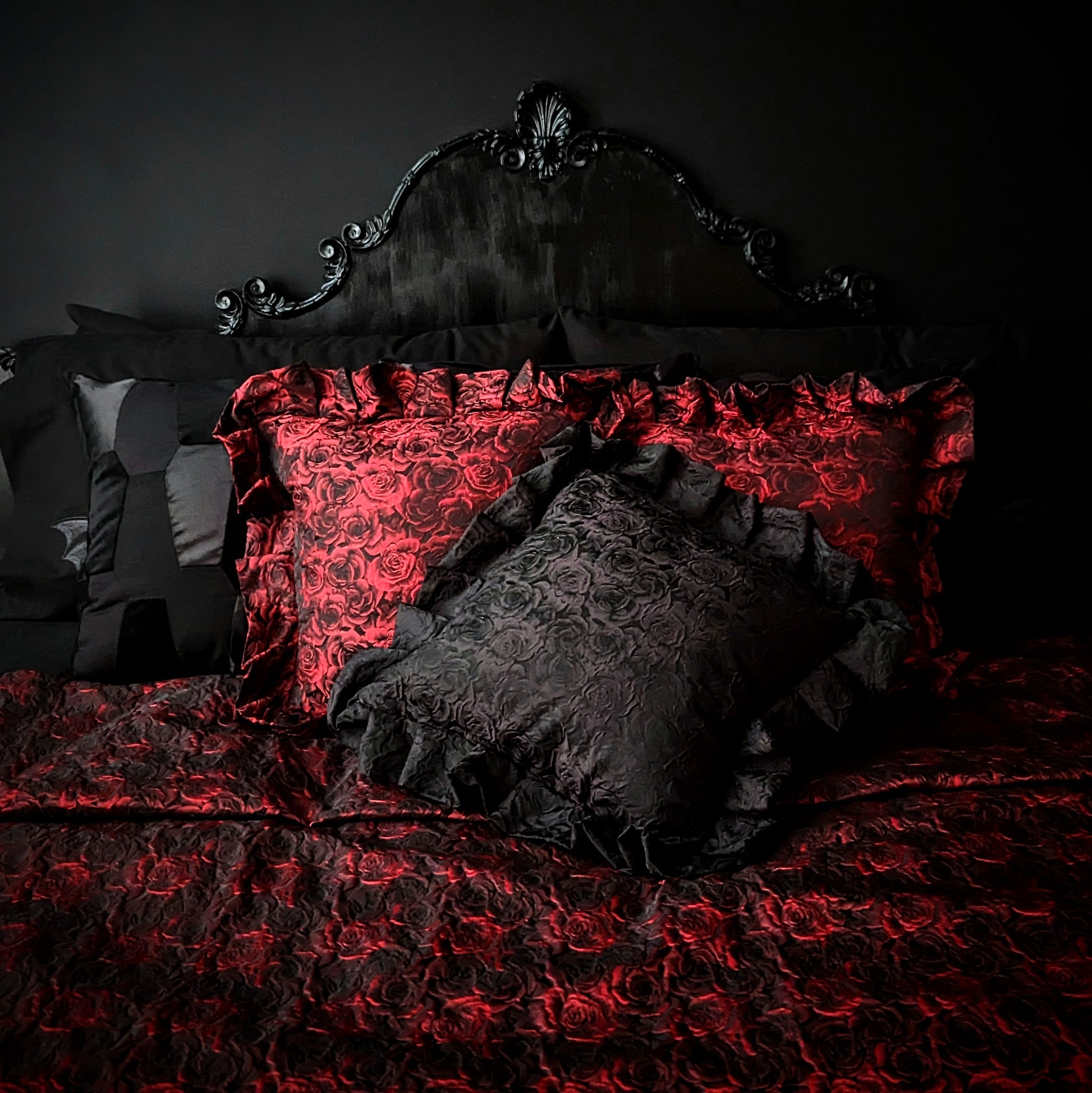 Black and red rose patterned cushions with ruffles around the edge on top of a matching throw in front of a black baroque headboard and black wall.