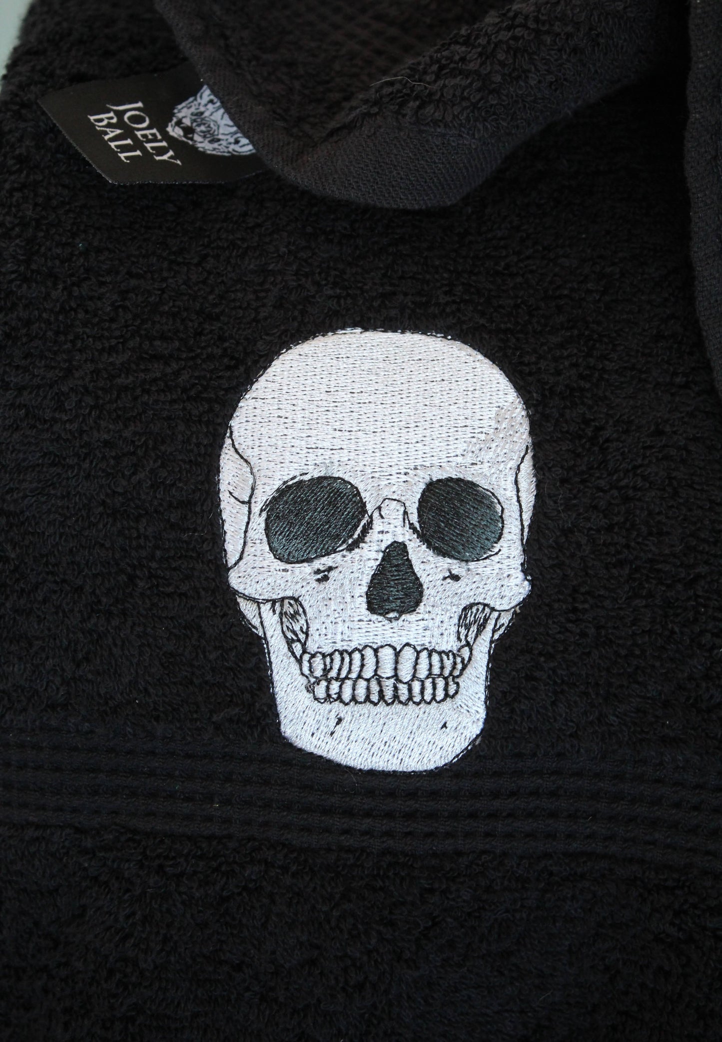 Detail shot of an anatomical skull embroidered on a black towel showing a Joely Ball label in the background.