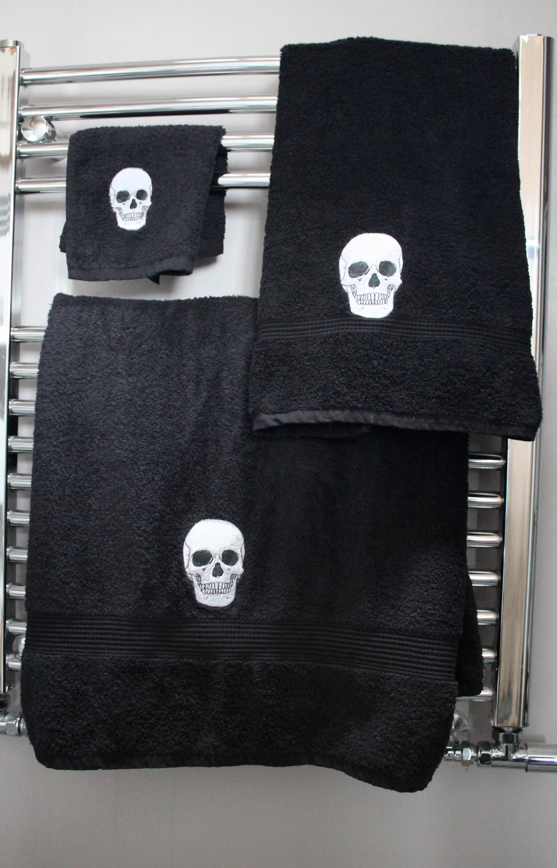 Black bath towel, hand towel and face cloth embroidered with anatomical skulls hanging on a chrome towel radiator.