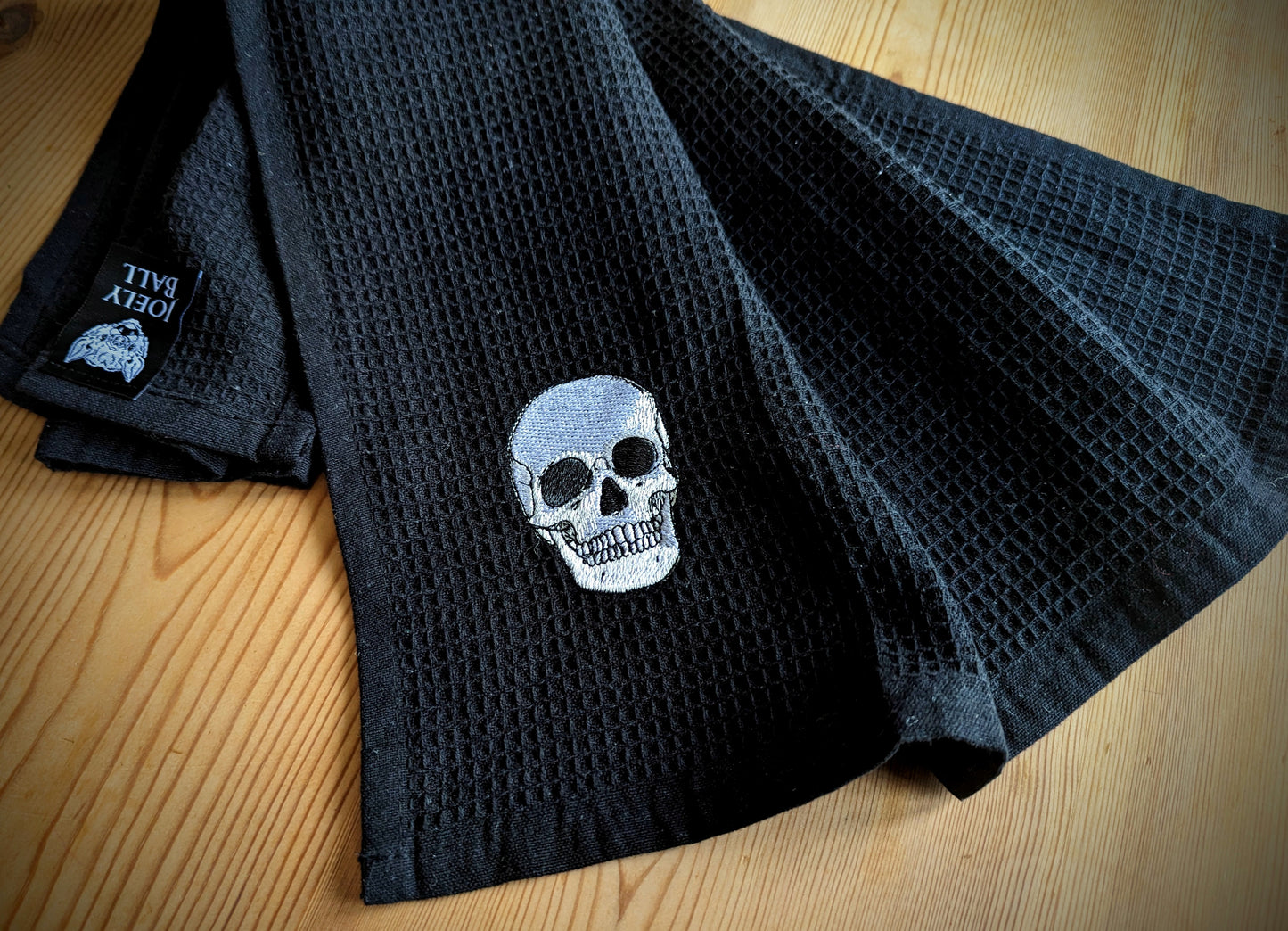 A black cotton waffle tea towel with an anatomical skull embroidered in the corner placed on a wooden surface .