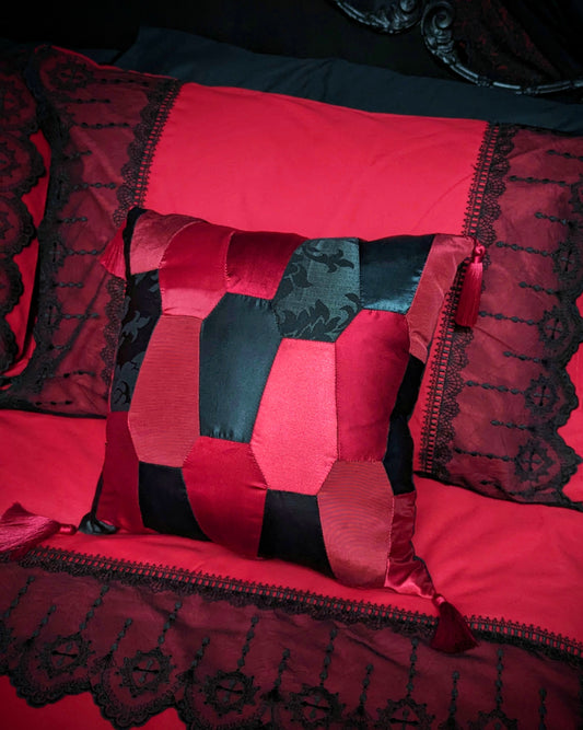 A red and black satin patchwork cushion with tassels on the corners, placed on a bed with red and black lace bedding and a black ornate backrest.