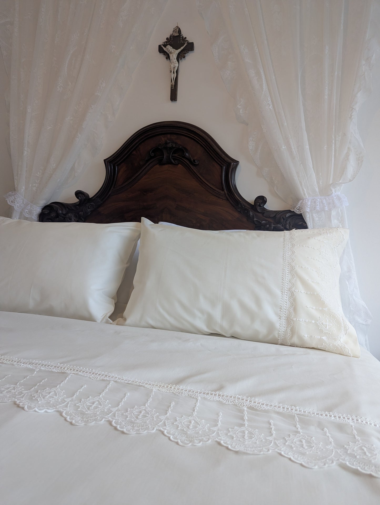 Bed with cream lace bedding, dark wood headboard and canopy, featuring a crucifix on the wall.