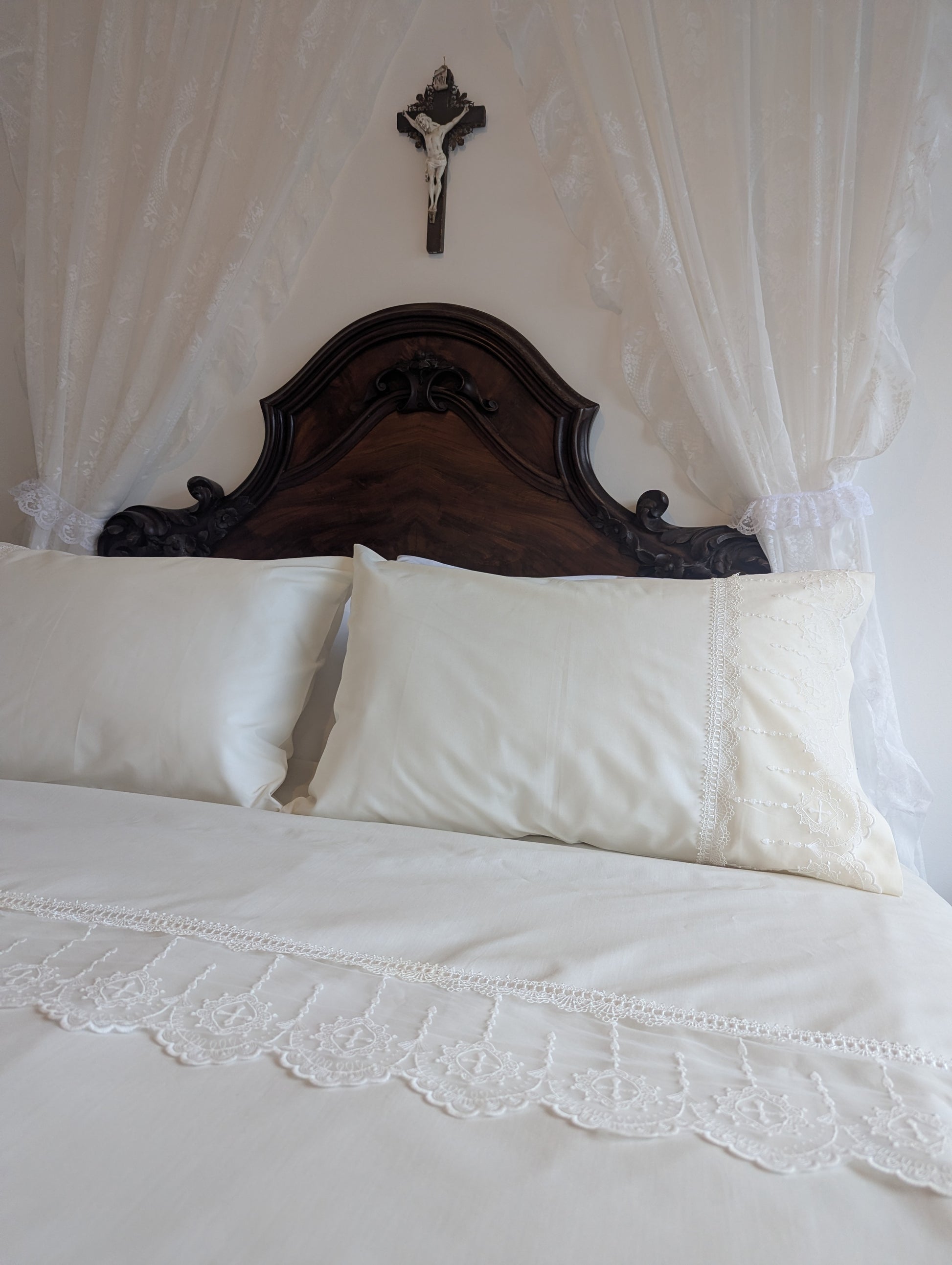 Bed with cream lace bedding, dark wood headboard and canopy, featuring a crucifix on the wall.